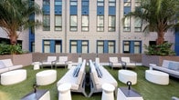 The 2nd Floor Courtyard is a thoughtfully designed amenity space that combines an expansive outdoor lounge with a fully equipped fitness center. The outdoor courtyard features artificial turf underfoot and is furnished with sleek, modern lounge beds, armchairs, and round side tables in a crisp white palette accented with blue-striped throw pillows. The space is framed by the surrounding multi-story building facade, with blue-trimmed windows and mature palm trees adding a resort-like ambiance. The fitness center is bright and airy, benefiting from abundant natural light streaming through large sliding glass doors and windows that overlook the courtyard. Floor-to-ceiling mirrors line the walls, visually expanding the space and enhancing the open feel. The gym is well-appointed with a full range of cardio equipment including treadmills and stationary bikes, a comprehensive dumbbell rack, cable and weight machines, a medicine ball rack, and a multi-purpose adjustable bench. Warm wood-toned flooring in a herringbone pattern adds a refined touch throughout. Built-in open shelving displays neatly rolled towels, plants, and motivational accents, while a ceiling fan ensures comfortable air circulation. A wall-mounted TV provides entertainment during workouts, completing this functional and stylish amenity offering.