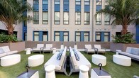 The 2nd Floor Courtyard is a thoughtfully designed amenity space that combines an expansive outdoor lounge with a fully equipped fitness center. The outdoor courtyard features artificial turf underfoot and is furnished with sleek, modern lounge beds, armchairs, and round side tables in a crisp white palette accented with blue-striped throw pillows. The space is framed by the surrounding multi-story building facade, with blue-trimmed windows and mature palm trees adding a resort-like ambiance. The fitness center is bright and airy, benefiting from abundant natural light streaming through large sliding glass doors and windows that overlook the courtyard. Floor-to-ceiling mirrors line the walls, visually expanding the space and enhancing the open feel. The gym is well-appointed with a full range of cardio equipment including treadmills and stationary bikes, a comprehensive dumbbell rack, cable and weight machines, a medicine ball rack, and a multi-purpose adjustable bench. Warm wood-toned flooring in a herringbone pattern adds a refined touch throughout. Built-in open shelving displays neatly rolled towels, plants, and motivational accents, while a ceiling fan ensures comfortable air circulation. A wall-mounted TV provides entertainment during workouts, completing this functional and stylish amenity offering.
