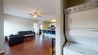 This single-floor unit offers a well-organized layout that connects living, dining, and kitchen spaces in an open, functional arrangement. The living area features rich hardwood flooring, a ceiling fan, and large windows fitted with blinds that allow generous natural light while providing privacy. Black leather seating and a central coffee table furnish the space, with views of greenery visible through the windows.

The kitchen is equipped with stainless steel appliances, including a refrigerator, electric range, and over-the-range microwave. White cabinetry with a mosaic tile backsplash and light countertops provide a clean, practical workspace. A breakfast bar with bar-height seating connects the kitchen to the dining area.

Each bedroom continues the hardwood flooring and features a built-in desk workspace, a closet with open shelving, and a window that brings in natural light. The rooms maintain a neutral, understated palette suited for functional living.

Two full bathrooms serve the unit, each featuring a tiled tub-shower combination, toilet, single-sink vanity with a white cabinet base, and a window for ventilation and light. Tile flooring completes each bathroom.

A stacked washer and dryer unit is conveniently located within the unit, adding in-home laundry capability.