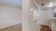 This single-story space showcases a practical and straightforward layout with notable architectural details that define its character. A defining feature is the pass-through window built into a dividing wall, complete with a stone or granite ledge, which connects two separate areas of the space — likely a living area and a kitchen or hallway. This design element promotes an open feel while maintaining distinct zones within the layout.

The flooring varies between rooms: soft, neutral-toned carpeting covers the more expansive open area to the left, while rich hardwood flooring extends through the adjacent hallway and connecting spaces. The walls throughout are finished in a clean, bright white, contributing to an airy and well-lit atmosphere. Vaulted or angled ceiling lines are visible in the carpeted room, adding a sense of height and architectural interest.

A hallway leads toward what appears to be additional rooms or exits, with a white door visible at the far end. Ceiling vents indicate a central HVAC system. Natural light filters in through a window visible through the pass-through, further brightening the interior and enhancing the overall sense of openness within this efficiently designed space.