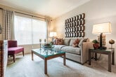 This single-floor residence offers a comfortable and cohesive layout designed for everyday living. The living room serves as the heart of the home, featuring plush carpeting, crown molding, a textured ceiling, and a large window dressed with patterned curtains that frame lush outdoor views, filling the space with natural light. Decorative touches, including a geometric mirror wall piece and coordinated accent pillows, lend the area a polished, welcoming ambiance.

The galley-style kitchen is efficiently organized with white cabinetry trimmed in warm wood tones, stainless steel appliances including a full-size refrigerator and dishwasher, and a double basin sink. Wood-look flooring and a woven area rug add texture and warmth to the space. Adjacent to the kitchen is a convenient in-unit laundry closet housing a full washer and dryer.

The primary bedroom is a serene retreat with soft carpeting, a walk-in closet with built-in shelving, and a private en-suite bathroom featuring an extended vanity with ample under-sink storage and a large mirror with vanity lighting. A second bedroom, also carpeted, includes a bright window with natural light and flexible use as a guest or flex room. A second full bathroom mirrors the first in its clean, functional design with wood-look flooring and a well-lit vanity.