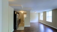 This single-floor space offers an open-concept layout that seamlessly connects the kitchen and living area, creating a practical and cohesive environment. The kitchen is equipped with stainless steel appliances, including a refrigerator and stove, complemented by dark cabinetry and light tile flooring that adds contrast to the space. A ceiling fan fixture provides both lighting and ventilation in the kitchen area. Adjacent to the kitchen, a washer and dryer area is conveniently tucked away, adding functional utility to the layout. The living area is distinguished by rich, dark hardwood flooring that extends throughout, creating a warm and polished aesthetic. Neutral beige walls provide a clean, versatile backdrop throughout the space. A window in the living area allows natural light to filter in, brightening the room and enhancing its open feel. Flush-mount ceiling fixtures provide consistent overhead lighting throughout. White baseboards offer a crisp finish that frames each room. The overall ambiance is straightforward and functional, with a neutral palette that lends itself well to a variety of interior styling preferences.