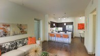 This single-floor space showcases a thoughtfully designed open-concept layout that seamlessly connects the living and kitchen areas. Rich, dark hardwood flooring runs throughout, providing a warm and cohesive foundation for the entire space.

The kitchen is a standout feature, outfitted with dark espresso cabinetry that extends to the upper walls, stainless steel appliances including a full-size refrigerator, and a microwave. A white peninsula counter serves as both a prep space and casual dining area, accommodating several orange bar stools that add a vibrant pop of color. Pendant lighting hangs above the peninsula, complementing the track lighting positioned along the kitchen ceiling to create a well-lit workspace.

The living area is open and inviting, with neutral-toned walls and ample room for comfortable furnishings. A sofa with decorative pillows anchors the space, while wall art adds personality. A doorway visible from the living area leads to an additional room, suggesting a practical and well-connected floor plan. The overall ambiance is modern and cohesive, balancing functional design with contemporary aesthetic appeal.