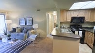 This single-floor residence is designed with an open-concept layout that seamlessly connects the kitchen and living area, creating a cohesive and functional living space. The kitchen is equipped with light wood cabinetry, a double-basin stainless steel sink, black appliances including a range and overhead microwave, and a peninsula countertop that serves as both a workspace and a natural divider between the kitchen and living area. The countertops feature a neutral, dark finish that complements the warm cabinetry tones.

The living area is carpeted and comfortably sized, offering enough room for a sofa, accent chair, and side tables. Soft green-toned walls run throughout the space, contributing to a calm and neutral ambiance. Decorative framed artwork adds visual interest to the living area walls.

A hallway visible from the main living space leads to additional rooms, suggesting a layout with private areas beyond. The flooring transitions from carpet in the living space to what appears to be a lighter surface in the hallway. Overall, the space presents a practical and well-organized interior with a cohesive, understated aesthetic suited for everyday living.