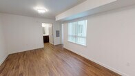 This single-floor residence at 21345–21359 International Blvd in Des Moines, Washington, offers a clean, modern interior with a cohesive design throughout. Warm hardwood-style flooring runs through the main living and kitchen areas, providing a consistent and inviting aesthetic that ties the space together.

The open living area is bright and simply appointed, with white walls, a large window fitted with vertical blinds that allow for adjustable natural light, and direct visual access to the bathroom. A separate carpeted room features both a window and a full glass exterior door with blinds, offering additional natural light and outdoor access — a practical and appealing feature.

The kitchen is well-equipped with dark espresso-toned cabinetry extending to the upper and lower walls, black appliances including a range, over-the-range microwave, and refrigerator, along with light countertops that contrast neatly against the cabinetry. A louvered interior door provides access to an adjacent space.

The bathroom continues the dark cabinetry theme with a matching vanity featuring multiple drawers and cabinet doors, a single sink with a large mirror above, and open wall shelving. Hardwood-style flooring extends into the bathroom, maintaining design continuity throughout the unit.