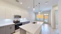 Located at 1345 14th St in Plano, TX, this single-floor, one-bedroom, one-bathroom unit offers a well-designed layout with modern finishes throughout. The kitchen is a standout feature, showcasing a large central island with white quartz countertops, an undermount sink, and sleek pendant lighting. Two-toned cabinetry — white uppers paired with gray lowers — frames stainless steel appliances, including a French door refrigerator, range, and built-in microwave, all set against a clean white tile backsplash. Light wood-style flooring runs through the main living and kitchen areas, adding warmth to the contemporary aesthetic. The bedroom features plush carpeting and a window fitted with blinds that allows for natural light and outdoor views. The bathroom continues the modern design with marble-look tile flooring, a dual-drawer vanity with white countertops, and a glass-enclosed walk-in shower with floor-to-ceiling marble-style tile and matte black hardware accents. A dedicated closet accessible from the bathroom provides organized storage with built-in shelving and hanging rods. The overall ambiance of the space is clean, bright, and cohesive, with a neutral color palette and contemporary detailing throughout.