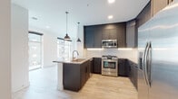 This single-floor residence presents a thoughtfully designed layout with contemporary finishes throughout. The kitchen serves as a central highlight, showcasing dark shaker-style cabinetry paired with granite countertops and a textured tile backsplash. An L-shaped peninsula with an undermount sink and pendant lighting above provides additional workspace and a natural gathering point. Stainless steel appliances, including a gas range, built-in microwave, and French door refrigerator, complete the kitchen's modern aesthetic. Under-cabinet lighting adds a refined touch, and light wood-toned flooring extends throughout the main living areas.

The living area is defined by expansive floor-to-ceiling windows that frame views of the surrounding cityscape, allowing abundant natural light to fill the space. A ceiling fan adds functional comfort. Bedroom spaces feature plush carpeting and large windows with outdoor views, creating a calm, bright atmosphere.

Two bathrooms serve the unit: one with a soaking tub and tiled shower surround with a built-in niche, and another with a glass-enclosed walk-in shower and large-format tile. Both bathrooms feature granite-topped dark vanities with light wood-look flooring, maintaining a consistent, cohesive design language throughout the residence.