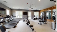 This fitness center is designed to accommodate a comprehensive workout experience within a thoughtfully organized single-level space. The layout is divided into distinct functional zones, allowing users to move seamlessly between cardio and strength training areas. The cardio section is outfitted with multiple Matrix treadmills, stationary and spin bikes, elliptical trainers, and a rowing machine, providing a broad range of aerobic options. A dedicated treadmill alcove features three side-by-side units positioned beneath a wall-mounted flat-screen television, flanked by windows that bring in natural light and outdoor views.

The strength training area is anchored by a multi-station cable machine, a free weight rack stocked with dumbbells, and adjustable benches. Large mirrors line the walls in this zone, enhanced by a striking rustic wood plank accent wall that adds warmth and visual contrast to the otherwise neutral gray and white palette. Recessed lighting throughout ensures consistent illumination, while a ceiling fan in the main area contributes to air circulation. Decorative artwork and a large wall clock add subtle character, creating a fitness environment that is both functional and visually inviting.