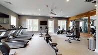 This fitness center is designed to accommodate a comprehensive workout experience within a thoughtfully organized single-level space. The layout is divided into distinct functional zones, allowing users to move seamlessly between cardio and strength training areas. The cardio section is outfitted with multiple Matrix treadmills, stationary and spin bikes, elliptical trainers, and a rowing machine, providing a broad range of aerobic options. A dedicated treadmill alcove features three side-by-side units positioned beneath a wall-mounted flat-screen television, flanked by windows that bring in natural light and outdoor views.

The strength training area is anchored by a multi-station cable machine, a free weight rack stocked with dumbbells, and adjustable benches. Large mirrors line the walls in this zone, enhanced by a striking rustic wood plank accent wall that adds warmth and visual contrast to the otherwise neutral gray and white palette. Recessed lighting throughout ensures consistent illumination, while a ceiling fan in the main area contributes to air circulation. Decorative artwork and a large wall clock add subtle character, creating a fitness environment that is both functional and visually inviting.