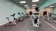 This fitness center is a comprehensively outfitted single-floor exercise space designed to accommodate a wide range of workout routines. The room features a distinctive color scheme, with teal accent walls contrasting against neutral beige tones, creating a clean and energizing atmosphere. A drop tile ceiling houses recessed fluorescent panel lighting, providing consistent, even illumination throughout the space.

The cardio section includes multiple treadmills by True Fitness, elliptical trainers, and stationary bikes, all arranged to allow comfortable spacing between users. The strength training area is equipped with a Paramount cable machine system, a leg press unit, weight benches, and a dedicated dumbbell rack with a full set of free weights. A wall-length mirror along one side of the room visually expands the space and allows users to monitor their form during exercises.

The flooring throughout consists of durable, dark speckled rubber matting, ideal for both equipment placement and user safety. A flat-screen television mounted near the ceiling provides entertainment for cardio users, and a water fountain is accessible along the mirrored wall. The space is accessed through a single keyed entry door with emergency exit signage, ensuring both security and safety compliance.