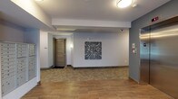 The lobby at Meridia Transit Crossing presents a well-appointed, single-floor common area designed to welcome residents and visitors alike. The entrance features full-glass double doors framed in dark metal, flanked by windows that allow natural light to fill the space. A textured stacked-stone accent wall adds architectural character near the entry, complementing the warm wood-look tile flooring that extends throughout.

The reception area is furnished with tasteful seating, including a cushioned armchair and a rustic wooden storage bench accented with decorative pillows, creating an inviting atmosphere. Metallic wall sculptures and curated artwork, including a bold abstract piece near the elevator corridor, contribute to the refined aesthetic. Soft blue-gray walls provide a cohesive, calming backdrop throughout.

Moving deeper into the lobby, residents have access to a bank of individual mailboxes conveniently located along one wall. A stainless steel elevator provides access to upper floors, positioned adjacent to the stone accent wall. The corridor leading to residential units features patterned carpet tiles, distinguishing it from the tiled lobby floor. Overhead, flush-mount ceiling fixtures provide consistent, even lighting. The overall design balances functionality with contemporary style, offering a comfortable and practical transitional space for daily resident use.