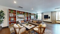 Located at 4121 Concord Creek Place in Richmond, VA, this well-appointed community lounge offers a welcoming and thoughtfully designed space for residents to gather and unwind. The open, single-floor layout features warm hardwood-style flooring throughout, creating a cohesive and inviting atmosphere.

The lounge area is anchored by a striking floor-to-ceiling built-in shelving unit finished in a bold terracotta red, displaying an eclectic mix of decorative objects, trailing plants, and curated artwork. Multiple seating arrangements — including plush sofas, armchairs, and accent chairs — are centered around a marble-topped coffee table atop a richly patterned area rug, fostering comfortable conversation and relaxation. A wall-mounted flat-screen television adds a media-friendly element to the space.

Adjacent to the lounge is a dedicated dining area featuring a sleek marble dining table surrounded by contemporary chairs. White cabinetry with granite countertops lines one wall beneath a vibrant red accent wall adorned with landscape artwork. Large windows flood this area with natural light and offer elevated views of the surrounding neighborhood.

Near the elevator vestibule, a secondary lounge nook with modern furniture provides additional seating, highlighted by warm orange accent walls and recessed lighting. The overall design blends bold color, natural textures, and modern furnishings to create an energetic yet comfortable communal environment.