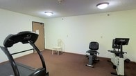 This single-floor fitness room offers a straightforward, functional space designed for basic exercise needs. The room features dark brown carpeting throughout, providing a practical surface underfoot, while the light-colored walls create a clean, neutral backdrop. Ceiling-mounted flush lighting illuminates the space evenly, ensuring visibility across the entire room.

The fitness area is equipped with a treadmill positioned near the entrance and a stationary recumbent bike toward the opposite side of the room, offering users both cardio and low-impact exercise options. A simple chair is also present, providing a place to rest between workouts. The room's layout allows for comfortable movement between equipment without feeling cramped.

A wood-paneled door provides access to the space, and electrical outlets are conveniently placed along the walls. The textured ceiling adds a subtle architectural detail consistent with the room's practical design. Overall, the fitness room presents a no-frills environment focused on functionality, suitable for light to moderate daily exercise routines.