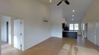 This single-floor space is defined by its striking vaulted ceilings, which lend an airy, expansive quality to the open-plan interior. The main living area is finished with light hardwood flooring that reflects natural light and contributes to the bright, clean aesthetic throughout. Crisp white walls are kept intentionally minimal, allowing the architectural volume of the space to take center stage. A ceiling fan is centrally mounted, adding both function and a subtle design element overhead, while recessed lighting provides consistent, even illumination across the room. A wall-mounted mini-split unit offers efficient climate control without disrupting the streamlined design. The kitchen, positioned toward the rear, features dark cabinetry that creates a bold contrast against the otherwise light palette. A peninsula-style counter provides workspace and casual seating potential, and a window above the sink brings in natural light. The overall ambiance is modern and unfussy, with a cohesive design language that balances practicality with a clean, contemporary aesthetic throughout the entire space.