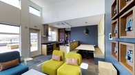 This single-floor space showcases a thoughtfully designed open-plan layout that seamlessly integrates a kitchen, dining area, and lounge zone. The interior is characterized by soaring ceilings with clerestory windows that draw in abundant natural light, creating an airy and expansive atmosphere. The kitchen is well-appointed with dark countertops, warm wood cabinetry, stainless steel appliances including a range with an overhead hood, and a central island that doubles as a prep and gathering space. Track lighting runs along the ceiling, complementing a striking black pendant light that anchors the dining area. The dining nook features a built-in booth with a yellow upholstered banquette set against a deep navy accent wall, providing a cozy yet modern setting. Two bold chartreuse accent chairs with blush pink cushions define the lounge area, adding a vibrant pop of color to the neutral palette. Flooring consists of a patterned area rug over what appears to be a hard surface floor, contributing texture and visual interest. A glazed door on one side offers outdoor access and additional natural light, enhancing the overall brightness and connectivity of the space.