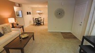 This single-floor space offers an open, flowing layout that seamlessly connects the living and dining areas. The flooring transitions between speckled terrazzo-style tile in the foreground and plush carpet in the dining area, adding visual variety and defining each zone. A raised wood-finished step subtly separates areas, contributing to the spatial definition without interrupting the open feel.

The dining area is anchored by a round dark-wood table surrounded by matching chairs, illuminated by a contemporary chandelier that draws the eye upward. A large circular decorative mirror mounted on the wall adds depth and a focal point to the dining space.

The living area features warm beige walls and recessed lighting that complement the natural tones of the flooring. Decorative wall art and a circular sculptural accent piece add character to the neutral palette. White baseboards frame the room cleanly throughout.

Overall, the space presents a warm, inviting ambiance with consistent neutral tones, thoughtful decorative touches, and a practical open-plan design that promotes easy movement and a cohesive aesthetic between living and dining functions.