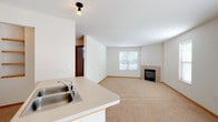 This single-floor unit features a bright, open-concept layout that seamlessly connects the kitchen and living areas. The kitchen is outfitted with white cabinetry, a white electric range, a full-size refrigerator, and a peninsula with a double-basin sink, providing functional workspace and a natural boundary between the kitchen and the carpeted living area. Track lighting and flush-mount fixtures illuminate the space effectively.

The spacious living room benefits from multiple windows that bring in natural light and offer outdoor views. A tile-surround fireplace serves as a focal point along one wall, adding a cozy architectural element. A wall-mounted PTAC heating and cooling unit ensures year-round climate control.

The bedroom is a well-proportioned, carpeted room featuring a window with blinds and a baseboard heating unit positioned beneath it. The bathroom includes tile flooring, a white vanity with an integrated sink, ample cabinet storage, and a large mirror. Warm wood-tone doors and trim are consistent throughout the unit, complementing the neutral color palette of white walls and beige carpet. The overall design is clean, functional, and straightforward.