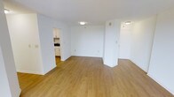 This single-floor space offers a clean and functional layout designed for comfortable everyday living. The main living area is generously proportioned, featuring warm light hardwood-style flooring that extends throughout, creating a cohesive and inviting atmosphere. Crisp white walls and flush ceiling lighting keep the space feeling bright and airy, while the open floor plan allows for flexible furniture arrangements to suit a variety of lifestyles.

A structural column adds a subtle architectural detail to the living area, defining the space without disrupting its openness. Adjacent to the main room, the kitchen is accessible through an open passageway and is outfitted with white cabinetry, a sink, and dark countertops — offering a tidy, efficient workspace. The white cabinetry contrasts neatly with the countertops, providing both style and practical storage.

Additional doorways visible from the main area suggest access to further rooms or storage spaces, contributing to the unit's overall sense of utility. The neutral color palette throughout — white walls, white trim, and light flooring — creates a versatile backdrop that accommodates a wide range of interior design preferences. The overall ambiance is clean, understated, and move-in ready.