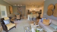 This single-floor residence showcases a thoughtfully designed open-concept layout that integrates the kitchen, dining, and living areas into one cohesive and functional space. The kitchen is well-appointed with stainless steel appliances, including a refrigerator and range, complemented by white cabinetry and a tiled backsplash. Pendant lighting in warm tones hangs above the kitchen island and counter, which features bar-height seating — ideal for casual dining or entertaining. Track lighting runs throughout the ceiling, providing consistent illumination across the space. The dining area is defined by a dark-toned table surrounded by upholstered chairs in a warm neutral hue, anchored beneath a bold typographic wall art piece that adds a contemporary artistic accent. The living area is carpeted and furnished with a plush sofa adorned with decorative pillows, paired with a coffee table resting on a patterned area rug that adds texture and visual interest. Crown molding along the upper walls adds an elegant architectural detail, while the overall color palette of soft grays, creams, and warm wood tones creates a welcoming and modern ambiance throughout.