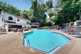 Located at 1901 Briarcliff Rd NE in Atlanta, Georgia, this outdoor amenity space offers a well-appointed pool and recreation area nestled within a lush, wooded setting. The centerpiece is a generously sized, geometric pool with clear turquoise water, accessible via entry steps with handrails and clearly marked depth indicators. Surrounding the pool is a wide concrete sun deck furnished with multiple lounge chairs, providing ample space for relaxation and sunbathing.

A tiered retaining wall constructed of weathered timber beams frames the rear of the pool area, creating a natural, layered landscape that rises into a dense canopy of mature hardwood trees. Colorful flowering plants and manicured shrubs are positioned throughout the deck and along the retaining walls, adding vibrant seasonal color to the environment.

An upper-level deck, accessible from the tiered landscaping, features outdoor dining furniture and a shaded umbrella table, offering a more intimate retreat within the wooded surroundings. String lights are visible along the fence line, suggesting a welcoming atmosphere during evening hours.

Adjacent to the pool is a single-story clubhouse structure with large windows and a modern design, providing convenient access to additional facilities. The entire space is enclosed by a wrought iron fence, ensuring privacy and security while maintaining open views to the surrounding natural landscape.
