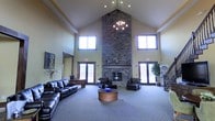 This single-floor space makes a striking first impression with its dramatic vaulted ceilings that rise to a sharp peak, creating an expansive and airy atmosphere. The focal point of the main lounge area is a towering floor-to-ceiling stacked stone fireplace, which anchors the room with a sense of warmth and grandeur. A classic chandelier hangs above, complementing the fireplace's rustic elegance with a touch of refined charm. Natural light pours in through multiple large windows positioned high along the walls and flanking the entry doors on either side, brightening the neutral-toned interior throughout the day. The lounge is furnished with rich, dark leather seating arranged around a central circular coffee table, offering a comfortable and inviting gathering area. Plush carpeting covers the main floor, adding warmth underfoot. French-style glass doors on both sides of the fireplace wall provide access points and contribute to the open, welcoming feel of the space. A large decorative mirror and tasteful wall art add personality to the room, while a potted tree near the entrance brings a natural, organic element indoors. Overall, the space combines rustic architectural character with comfortable, functional furnishings to create an inviting communal environment.