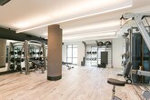Located in Mountain View, CA, this modern fitness center offers a well-appointed, single-floor workout facility designed with both functionality and aesthetics in mind. The space features light-toned hardwood flooring accented with subtle patterned inlays, creating a clean and inviting atmosphere throughout.

The facility is equipped with a comprehensive selection of professional-grade exercise machines, including cable and weight machines by Hoist, a full dumbbell rack with a wide range of weights, and an adjustable workout bench. Additional free weights and functional training equipment such as kettlebells and medicine balls are neatly organized along the walls.

Architectural highlights include floor-to-ceiling black-framed glass windows and walls, which flood the interior with abundant natural light while offering open views of the surrounding neighborhood and landscaping. Linear LED lighting runs across the ceiling, providing bright, even illumination throughout the space. A structural column anchors the center of the room, adding an industrial design element.

Full-length mirrors line one wall of the strength training area, enhancing the sense of space and supporting proper form during workouts. A mounted flat-screen display and a built-in open shelving unit round out the amenities, contributing to both the utility and modern character of the facility. The overall ambiance is clean, bright, and purpose-driven.