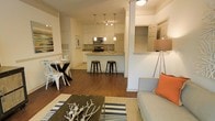 This single-floor residence is designed around an open-concept layout that integrates the living, dining, and kitchen areas into one cohesive, flowing space. Warm hardwood flooring runs throughout, lending continuity and a sense of warmth to the entire floor plan.

The kitchen serves as a functional centerpiece, featuring white cabinetry, granite or stone countertops, a tile backsplash, and stainless steel appliances including a range and microwave. A peninsula with bar-height seating accommodates two black metal stools, creating an ideal spot for casual dining or conversation. Pendant lighting with multiple adjustable fixtures hangs above the peninsula, providing focused task lighting.

Adjacent to the kitchen, a compact dining area fits a round glass-top table with metal chairs, maximizing space efficiency without sacrificing style. The living area is furnished with a neutral upholstered sofa accented by an orange throw pillow, a striped area rug, and a rustic driftwood coffee table, contributing to the space's relaxed coastal character.

Structural columns and partial walls define zones without enclosing them, maintaining the open, airy feel throughout. The overall ambiance is bright, casual, and well-organized.