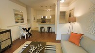 This single-floor residence is designed around an open-concept layout that integrates the living, dining, and kitchen areas into one cohesive, flowing space. Warm hardwood flooring runs throughout, lending continuity and a sense of warmth to the entire floor plan.

The kitchen serves as a functional centerpiece, featuring white cabinetry, granite or stone countertops, a tile backsplash, and stainless steel appliances including a range and microwave. A peninsula with bar-height seating accommodates two black metal stools, creating an ideal spot for casual dining or conversation. Pendant lighting with multiple adjustable fixtures hangs above the peninsula, providing focused task lighting.

Adjacent to the kitchen, a compact dining area fits a round glass-top table with metal chairs, maximizing space efficiency without sacrificing style. The living area is furnished with a neutral upholstered sofa accented by an orange throw pillow, a striped area rug, and a rustic driftwood coffee table, contributing to the space's relaxed coastal character.

Structural columns and partial walls define zones without enclosing them, maintaining the open, airy feel throughout. The overall ambiance is bright, casual, and well-organized.