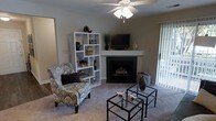 This single-floor space offers a warm and inviting atmosphere anchored by a central fireplace with a clean mantel, perfect for both relaxing and entertaining. The living area is finished with wall-to-wall carpeting that adds comfort underfoot, while neutral-toned walls create a versatile backdrop for a variety of décor styles. A ceiling fan provides both lighting and airflow, contributing to year-round comfort. Natural light filters in through a sliding glass door fitted with vertical blinds, hinting at outdoor access and brightening the interior. The layout accommodates a thoughtful arrangement of seating, including a patterned accent chair, alongside modern nesting glass-top coffee tables that add a contemporary touch. A modular open-shelving unit provides both display and storage functionality, enhancing the room's organization and style. The mantel above the fireplace serves as a natural focal point, offering space for decorative elements. Overall, the space balances practicality with aesthetic appeal, making it a comfortable and functional single-level home.