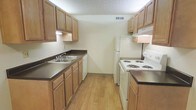 This single-story space is designed with practicality and functionality in mind. The kitchen serves as a central highlight, laid out in a classic galley configuration that maximizes efficiency. Light wood cabinetry lines both sides of the kitchen, providing generous storage space both above and below the countertops. The dark laminate countertops offer a clean contrast to the warm tones of the cabinetry, creating a visually cohesive look. A double-basin sink is positioned conveniently along one wall, accompanied by a dishwasher for added convenience. On the opposite side, a white electric coil range and a full-size refrigerator complete the appliance suite, keeping the kitchen well-equipped for everyday use. An overhead range hood is installed above the stove, ensuring proper ventilation. The kitchen flooring features light wood-tone laminate, adding warmth to the space. A textured ceiling and a centrally placed ventilation duct reflect a practical, no-frills interior design approach. Under-cabinet lighting provides functional illumination along the workspace. The layout is straightforward and well-organized, making it an accessible and efficient environment for daily use.