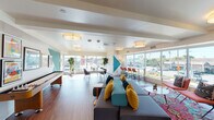 Located at 515 W Colorado St in Glendale, California, this single-floor community lounge is a thoughtfully designed social and collaborative space that blends modern comfort with bold, energetic aesthetics. The open-plan layout is unified by warm hardwood flooring and flush-mount drum lighting throughout, creating a bright and welcoming atmosphere.

The lounge is defined by expansive floor-to-ceiling windows along the street-facing walls, flooding the interior with abundant natural light and offering open views of the surrounding Glendale neighborhood. A striking geometric mural in teal, yellow, and red serves as a focal point on one wall, lending the space a lively, contemporary character.

The seating area features a large sectional sofa accented with teal and yellow throw pillows, complemented by a vibrant patterned area rug and accent chairs. A wall-mounted flat-screen television anchors the entertainment zone, while a set of private high-back booth seating provides a more intimate gathering option.

A bar-height counter with red stools runs along the window line, ideal for casual seating with street views. The space also includes a dedicated collaborative work area with a long communal table and modern chairs, as well as a shuffleboard table for recreation. Curated artwork and potted plants are placed throughout, adding warmth and a polished finishing touch to this dynamic, multi-use community space.