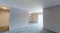This single-floor space presents an open and airy layout characterized by neutral gray and beige wall tones that contribute to a calm, understated ambiance. The living area is finished with wall-to-wall carpet in a soft gray hue, providing a comfortable and cohesive foundation throughout the space. Smooth white ceilings at a standard height enhance the sense of openness, while clean white baseboards offer a crisp contrast to the neutral walls.

A notable feature is the sliding glass door fitted with vertical blinds, which allows generous natural light to filter into the interior and provides a direct connection to the outdoors. This element brightens the space considerably, making it feel welcoming and open. A modest chandelier suspended from the ceiling in the adjacent area serves as a subtle focal point, adding a touch of understated elegance to the room.

The layout appears to flow openly between defined areas, offering flexibility in how the space can be arranged and utilized. Electrical outlets and a wall vent are discreetly placed, maintaining the clean visual lines of the interior. Overall, the space conveys a simple, well-maintained character suited to a variety of functional arrangements.