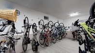 Located at Breakwater, this dedicated bicycle storage room is designed to keep residents' bikes organized and readily accessible. The space features a clean, minimalist aesthetic with bright white walls and smooth light-toned flooring that together create a tidy, well-maintained environment. A row of individually numbered wall-mounted bicycle hooks lines the main wall, accommodating a variety of bike styles — from folding bikes and road bikes to mountain bikes and hybrid models — keeping each one neatly suspended and easy to retrieve. The numbered slots, running sequentially along the wall, ensure that each resident can quickly identify their designated space. Overhead lighting provides consistent, even illumination throughout the room, enhancing both visibility and security. The layout is thoughtfully arranged to maximize the number of bikes stored while still allowing comfortable movement through the space. A decorative wall sign adds a subtle touch of personality to an otherwise utilitarian room. Contained on a single floor, this bike storage facility reflects a commitment to practical, resident-focused amenities, making it an ideal resource for those who rely on cycling for commuting, recreation, or everyday errands.