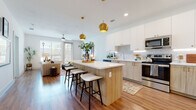This contemporary single-floor residence is designed with a clean, modern aesthetic that emphasizes open living and functional elegance. The heart of the home is a spacious open-concept kitchen and living area, where rich hardwood flooring flows seamlessly throughout. The chef's kitchen is a standout feature, showcasing a large center island with a white quartz countertop and natural wood cabinetry, comfortably accommodating bar seating. Upper white cabinetry contrasts beautifully with the warm wood lower cabinets, while a subway tile backsplash and stainless steel appliances — including a refrigerator, range, microwave, and dishwasher — complete the well-equipped space. Statement gold globe pendant lights add a sophisticated touch overhead. The adjacent living area is bright and inviting, benefiting from large windows and a glass door that welcome natural light and outdoor views, complemented by a ceiling fan. Two well-sized bedrooms offer comfort and tranquility, each featuring plush carpeting, ceiling fans, and generous windows. The bathroom is finished with warm hardwood-style flooring, a sleek vanity with wood cabinetry, and a full walk-in glass-enclosed shower, all set within a clean, bright layout that reflects the home's overall modern sensibility.