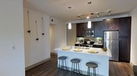 This single-floor unit showcases a thoughtfully designed open-concept layout, seamlessly connecting the kitchen and living areas for an airy, functional feel. The kitchen serves as a central focal point, featuring dark espresso cabinetry that creates a striking contrast against the crisp white quartz peninsula countertop. A trio of bar stools lines the peninsula, providing casual seating and defining the boundary between kitchen and living space. Stainless steel appliances, including a refrigerator, stove, and built-in microwave, add a sleek, modern touch. A subway tile backsplash complements the cabinetry and contributes to the contemporary aesthetic. Overhead, a combination of track lighting and two elegant pendant lights illuminate the workspace effectively. The flooring throughout is a rich, dark hardwood, adding warmth and continuity across the open floor plan. The walls are finished in a clean, neutral white, enhancing the sense of brightness in the space. A hallway visible in the background suggests additional rooms beyond the main living area, adding to the functional layout of this well-appointed unit.
