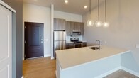 Located at 1455 W Lake St in Minneapolis, MN, this contemporary single-floor unit offers a thoughtfully designed layout with modern finishes throughout. The open-concept kitchen serves as a central hub, featuring stainless steel appliances including a refrigerator, gas range, and built-in microwave, complemented by rich wood-toned cabinetry, a classic subway tile backsplash, and a large island with a quartz countertop and undermount sink. Pendant globe lighting adds a stylish touch overhead, while light hardwood flooring anchors the space with warmth. The living area extends toward a private balcony, offering views of the surrounding Minneapolis neighborhood and filling the space with natural light. The carpeted bedroom is generously sized, with a large window framing treetop and cityscape views. A spacious walk-in closet provides extensive wire shelving and hanging storage. The bathroom is well-equipped with a corner soaking tub, subway-tiled shower with grab bar, dual-sink vanity with wood-framed mirror and dark cabinetry, and clean tile flooring. The unit presents a cohesive blend of modern design and functional living tailored for urban life.