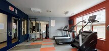 Located at 201 Railroad Ave in East Rutherford, NJ, this well-equipped fitness center offers a comprehensive workout environment across a single floor. The space is defined by bold, energizing design choices, including deep red accent walls paired with cool blue and gray tones, complemented by a vibrant checkerboard-patterned tile floor in orange and gray.

The cardio zone is generously stocked with treadmills, elliptical trainers, upright stationary bikes, and interactive cycling bikes equipped with individual display screens. Wall-mounted flat-screen TVs are positioned throughout for entertainment during workouts. Natural light flows in through multiple windows fitted with blinds, and large glass doors open to an outdoor area, enhancing the sense of openness.

A dedicated strength and functional training section features a multi-station cable machine, a full rack of dumbbells, medicine balls in varying weights, an exercise stability ball, and a freestanding punching bag. The glass-paneled entry doors with navy blue framing give the facility a polished, professional appearance and allow visibility into the space from the corridor.

The overall ambiance is motivating and functional, designed to support a full range of fitness activities in a clean, modern environment.