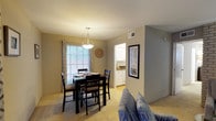 This single-floor residence offers a well-organized and inviting layout that combines character-rich design with everyday functionality. The living room serves as the heart of the home, anchored by a striking painted brick accent wall that adds warmth and texture to the space. Soft carpeting underfoot and neutral wall tones create a relaxed atmosphere, while the room comfortably accommodates a full seating arrangement centered around a glass-top coffee table. A wall-mounted flat-screen television and carefully placed lighting complete the setup, making it an ideal space for both relaxing and entertaining.

Adjacent to the living room, the dining area is bathed in natural light from a large window dressed with patterned curtains. A pendant light fixture hangs above the dining table, adding a refined focal point to the room. The space easily accommodates a four-seat dining set, providing a comfortable area for daily meals or hosting guests.

A glimpse through the dining area reveals a connected kitchen featuring white cabinetry and clean countertops, contributing to the home's bright and cohesive aesthetic. The open flow between the living, dining, and kitchen areas enhances the overall sense of space, making this residence feel open, airy, and thoughtfully designed throughout.