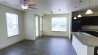 This single-floor space offers a clean, open layout designed for comfortable everyday living. The main living area is anchored by rich, dark hardwood-style flooring that runs throughout, creating a warm and cohesive aesthetic. A ceiling fan with a light fixture provides both comfort and illumination, while a set of track lights adds functional overhead lighting to the space.

Large windows dressed with horizontal blinds line the walls, flooding the interior with abundant natural light and offering views of the surrounding exterior. A glass-panel door provides additional access and light, enhancing the airy feel of the room.

The living area flows naturally toward the kitchen, which is partially visible and features pendant lighting hanging above what appears to be a counter or island with dark stone surfaces, complemented by white cabinetry below. The kitchen also includes dark appliances, adding a modern contrast to the light-toned walls.

Throughout the space, neutral beige walls and clean lines contribute to a bright, understated ambiance. The overall design is practical and inviting, with thoughtful details such as vaulted ceiling accents and well-placed lighting that enhance the sense of openness and scale across the entire floor plan.