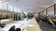 This health club is a spacious, single-floor fitness facility designed to accommodate a comprehensive range of workout routines. The layout is thoughtfully organized into distinct zones, creating an efficient and functional training environment. A generous cardio section lines one side of the main floor, housing a full row of treadmills, elliptical trainers, and stationary bikes — including recumbent and upright models — all equipped with individual digital displays. Opposite the cardio equipment, a well-stocked strength training area features multiple selectorized cable and resistance machines suitable for targeting various muscle groups. At the far end of the main floor, a dedicated free weights zone offers squat racks, weight benches, a padded mat area for floor work, and an assortment of dumbbells. A separate, mirror-lined room provides additional space for free weight exercises and stretching. The facility is finished with light-toned, easy-to-clean flooring throughout the main area and rubberized flooring in the free weights room for added safety. Sleek linear LED lighting runs across the drop-panel ceiling, while large clerestory windows along the upper walls admit abundant natural light and views of the surrounding urban streetscape. A wall-mounted television and digital check-in panel are also present, contributing to a modern, well-appointed atmosphere.