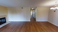This single-floor residence offers a straightforward and functional layout designed for comfortable everyday living. The main living area is generously open, featuring rich, warm-toned hardwood-style flooring that extends throughout the space, providing a cohesive and inviting feel underfoot. The walls are finished in a neutral off-white tone, creating a clean, bright backdrop that allows for versatile interior styling. A textured popcorn ceiling adds a classic residential character to the space, while flush-mounted ceiling light fixtures provide consistent overhead illumination throughout.

A central hallway extends from the main living area and leads to multiple doorways, suggesting several separate rooms — likely bedrooms and a bathroom — arranged along the corridor. The hallway is well-lit, maintaining the open and airy quality of the overall space. Ventilation is provided through a wall-mounted HVAC vent visible in the main area, ensuring climate control throughout. Clean white baseboards frame the flooring along every wall, adding a crisp finishing detail. The overall ambiance is practical and move-in ready, with a neutral palette and durable finishes that appeal to a wide range of occupants.