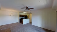 This single-floor residence features an open-concept layout that connects the living area and kitchen in one cohesive space. The living area is finished with wall-to-wall carpeting in a neutral gray tone, providing a comfortable and understated foundation. A ceiling fan with wooden blades is centrally mounted overhead, offering both functional air circulation and a classic design accent.

The kitchen is positioned at the rear of the open layout and is defined by dark wood cabinetry that extends to the upper walls, providing ample storage space. Black appliances, including a range and microwave, complement the cabinetry and contribute to a consistent, coordinated aesthetic. A raised peninsula separates the kitchen from the living area, creating a natural transition between the two spaces while also offering potential seating or serving space.

Overhead fluorescent lighting in the kitchen ensures a well-lit work area, while the neutral white walls throughout keep the space feeling open and bright. The single-floor layout promotes ease of movement and accessibility, making the overall floor plan practical and efficient for everyday living.