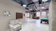 Located at 1 Gold St in downtown Hartford, CT, this generously sized game room offers a well-rounded recreational experience within an industrial-chic setting. The space is defined by its exposed ceiling with visible ductwork and conduit, accented by a series of pendant industrial lights that cast a warm, ambient glow across the light-toned herringbone tile flooring below.

The room is thoughtfully arranged to accommodate a variety of activities. A full-size pool table with a vibrant teal felt surface serves as a central focal point, flanked by a ping-pong table and a foosball table, making it ideal for social gatherings and competitive play. Comfortable leather sofas and accent chairs, including a patterned armchair and bold yellow seating, are arranged throughout the space to provide relaxed lounging areas.

Large windows along the perimeter flood the room with natural light and offer views of the surrounding Hartford cityscape, enhancing the open and airy feel of the space. A wall-mounted oversized Connect Four game adds a playful, interactive element to the décor. Additional seating with rolling task chairs suggests a flexible, multipurpose use. Structural concrete columns add to the industrial character of the room, contributing to its urban loft aesthetic. The overall ambiance is dynamic, inviting, and well-suited for community recreation.