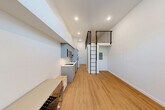 Located on University Way NE in Seattle's vibrant University District, this thoughtfully designed micro-unit maximizes every inch of its compact footprint through smart vertical planning and modern finishes. The main living area features warm-toned hardwood-style flooring throughout and soaring ceilings that lend an open, airy feel well beyond what the floor area suggests. A striking black metal ladder leads to an elevated loft level enclosed by a sleek horizontal-rail railing, providing an additional functional space overhead.

The open-plan layout seamlessly integrates the living, dining, and kitchen zones. The kitchenette is outfitted with gray flat-front cabinetry, a countertop cooktop, single-basin sink, dishwasher, and stainless steel microwave — all efficiently arranged along one wall. Recessed lighting illuminates the space, while a large window at the end of the unit fills the room with natural light and frames views of surrounding greenery.

A stacked washer and dryer unit is conveniently housed within the unit, adding practical value. The bathroom features a clean, contemporary design with a white vanity, toilet, and a glass-door walk-in shower, finished with neutral tile flooring. The overall aesthetic is minimalist and modern, with a crisp white palette that enhances the sense of space throughout.
