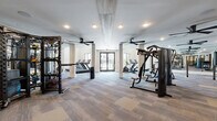 This expansive fitness center is thoughtfully designed to accommodate a wide range of workout routines within a single, open floor plan. The space is anchored by durable, multi-tone carpet tile flooring and illuminated by recessed ceiling lights complemented by modern black ceiling fans throughout, creating a comfortable and well-ventilated environment. Structural columns define distinct workout zones without interrupting the overall sense of openness.

The cardio section lines one side of the facility with a row of treadmills and elliptical machines, positioned near large windows and French doors that allow natural light to pour in and provide views to an outdoor area. Opposite this zone, a comprehensive strength training area features cable machines, a multi-station weight rack, and a full dumbbell and kettlebell rack arranged in front of floor-to-ceiling mirrors that visually expand the space. A dedicated boxing corner houses heavy punching bags suspended from the ceiling, adding a dynamic training option. Additional specialty equipment, including a stair climber and recumbent bike, rounds out the cardio offerings.

Toward the rear of the facility, glass-panel doors lead into a separate studio or ancillary room, ideal for group classes or focused training. A built-in water fountain station is conveniently located near the entry to this area. The overall aesthetic is modern and clean, with neutral tones, white walls, and dark equipment creating a sleek, professional atmosphere.
