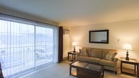 This single-floor residential space offers a welcoming and functional layout well-suited for everyday living. The living room serves as the centerpiece of the unit, featuring a large sliding glass door framed by vertical blinds and dark accent drapes, which floods the space with natural light and provides direct access to an outdoor area. A wall-mounted air conditioning unit is positioned above the sliding door, ensuring year-round comfort. The room is finished with neutral-toned carpeting underfoot and smooth, light-colored walls that enhance the sense of openness. Furnishings include a plush sofa accented with decorative pillows, a wood and glass coffee table, and a side table topped with a warm table lamp that adds an inviting glow to the space. A framed rectangular mirror mounted on the wall contributes to the room's depth and brightness. A baseboard heater runs along the lower wall, providing an additional source of warmth. The overall ambiance is calm and understated, with a neutral palette and practical design choices that create a livable, comfortable environment throughout.