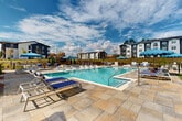 Located at 100 Oakbrook Drive in McDonough, Georgia, this outdoor amenity area centers around a generously sized resort-style swimming pool with crystal-clear turquoise water. The pool deck is finished in elegant stone tile pavers, providing ample space for lounging and entertaining. Numerous chaise lounges with navy blue cushions are arranged along the pool's perimeter, complemented by vibrant blue market umbrellas that offer shade throughout the day. A dedicated shallow sun shelf extends along one side of the pool, ideal for relaxation at the water's edge. Adjacent to the main pool, a separate spa provides an additional retreat. A handsome wood pergola anchors one corner of the deck and shelters an outdoor grilling and kitchen station, perfect for al fresco dining. Shaded seating areas with tables and chairs are scattered throughout the deck, offering flexible gathering spaces. A pool house structure provides convenient access to restroom facilities. Manicured evergreen topiaries and ornamental plantings add refined landscaping details, while a backdrop of tall Georgia pines and open sky enhances the natural ambiance. The surrounding multi-story residential buildings frame the community setting, and a fenced perimeter ensures privacy and safety throughout this well-designed outdoor living space.