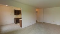 This single-floor residence offers a practical and functional layout designed for comfortable everyday living. The main living area is finished with neutral wall-to-wall carpeting, providing a warm and quiet atmosphere throughout the space. An open pass-through connects the living area to the kitchen, which features dark wood cabinetry, a countertop with an integrated sink, and tile flooring that contrasts neatly with the carpeted areas. The kitchen's darker cabinetry adds a grounding visual element to the otherwise light and neutral color palette found throughout the home. The walls are finished in a soft, muted tone that contributes to a clean and understated ambiance. A hallway visible from the main living area leads to additional spaces, including what appears to be a closet with built-in shelving, suggesting well-considered storage solutions. The open sight lines between the living and kitchen areas create a sense of connectivity, making the space feel cohesive and easy to navigate. Overall, the residence presents a straightforward, move-in-ready layout with a neutral aesthetic well-suited for a variety of furnishing styles.