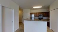 This single-floor residence offers a well-organized layout designed for comfortable, everyday living. The kitchen serves as the heart of the space, outfitted with rich, warm-toned wood cabinetry providing ample storage, granite countertops with natural stone patterning, and a full suite of stainless steel appliances including a refrigerator, electric range, and built-in microwave. A peninsula counter extends outward, creating a natural divide between the kitchen and the adjoining living area while offering additional prep and seating space. Overhead, a fluorescent light fixture ensures the kitchen is well-lit throughout the day. The flooring transitions from the kitchen area into soft, neutral-toned carpeting in the adjacent living spaces, contributing to a cozy and cohesive aesthetic throughout. The walls are finished in a clean, light neutral tone that reflects light and enhances the sense of openness. A hallway visible from the kitchen connects to additional rooms within the unit, suggesting a thoughtful and practical floor plan. The overall ambiance is understated and move-in ready, with functional finishes that balance utility and comfort across the entire space.