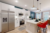 This single-floor unit is defined by a cohesive contemporary design with bold teal accent walls that run through the kitchen, living area, and bedroom, creating a vibrant visual thread throughout the space. The open-concept kitchen is a standout feature, outfitted with full-height white shaker cabinets, black hardware, a white quartz countertop, and a central island with bar seating. Stainless steel appliances—including a French door refrigerator, range, microwave, and dishwasher—are complemented by under-cabinet lighting and light hardwood-style flooring. The kitchen flows seamlessly into the living area, which benefits from large windows that draw in natural light and is anchored by a striking sputnik chandelier. The bedroom continues the teal accent theme, featuring plush carpeting, generous natural light from multiple windows, and a ceiling fan. A full bathroom adjacent to the bedroom offers a white vanity with quartz countertop, black fixtures, a framed mirror, and a tub-shower combination with marble-style tile surround. A dedicated laundry area accommodates a stacked washer and dryer unit alongside wire shelving for storage.